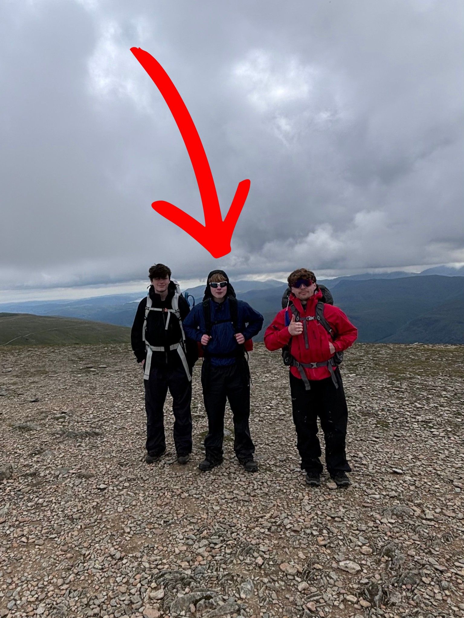 Euan, centre and marked with a red arrow, with two friends on a mountain summit, with hills and low cloud behind.